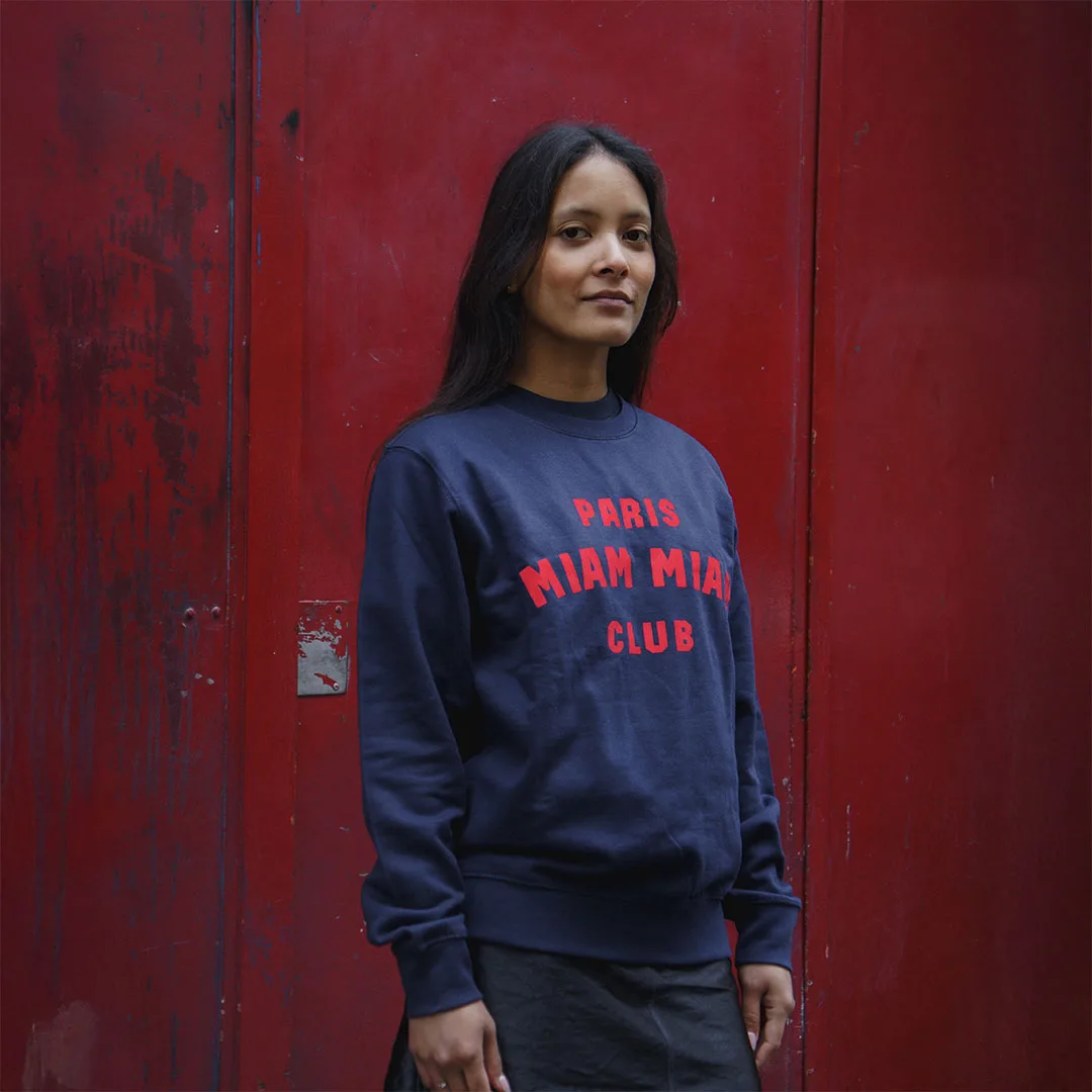 Model wearing a navy blue Paris Miam Miam Club sweatshirt in front of a red door