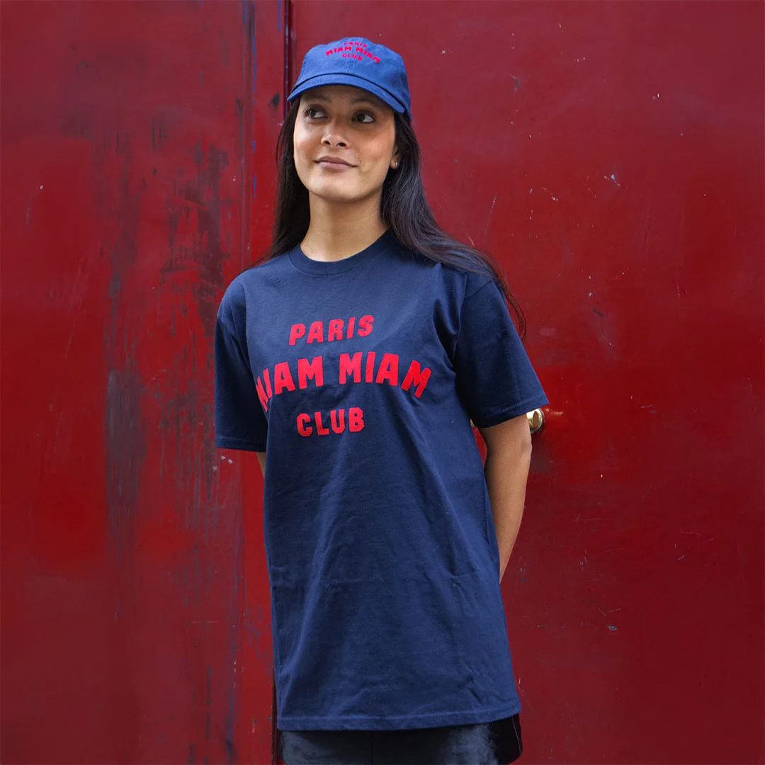 Model wearing a navy blue Paris Miam Miam Club T-shirt and cap in front of a red door