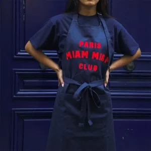 Close-up of a Paris Miam Miam Club apron worn over a navy blue T-shirt in front of a blue door